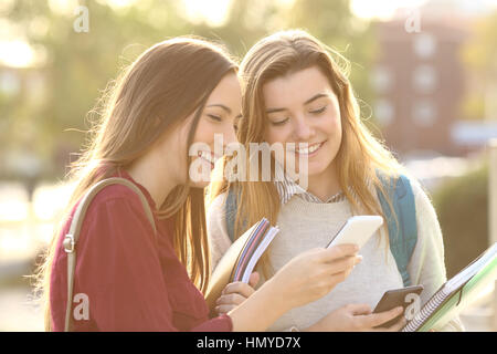 Two beautiful students watching media content on line in a smart phone outdoors in a park or university campus Stock Photo