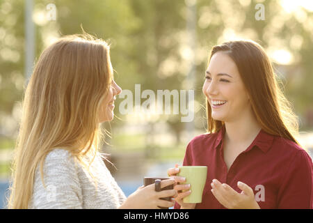 Two happy friends talking holding coffe mug outdoors in a park with a green background at sunset with a warm back light Stock Photo