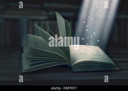 Opened magic book with magic light on a wooden table with bookshelv, invitation to study literatures. The magic of reading, storytelling and education Stock Photo