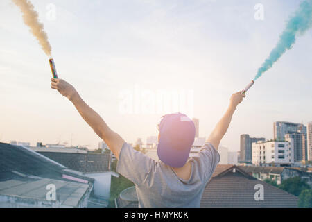 Male teenager holding colorful smoke sticks up in the air over urban city background Stock Photo