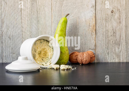Blue Cheese in a white ceramic recipient with a pear, three walnuts on a black table with weathered wood gray background Stock Photo