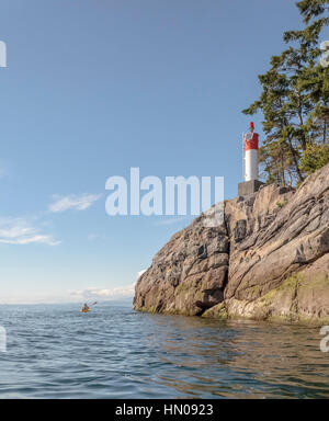 A 64 year old man is seen in the distance kayaking past a rocky headland with a lighthouse, on a sunny day off the Sunshine Coast, British Columbia. Stock Photo
