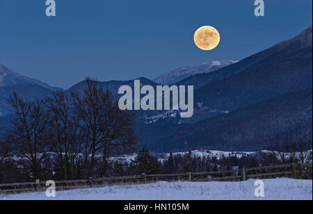 Enchanting nocturnal rural scene with full moon rising over the Piatra Mare (Big Rock) peak as seen from Fundata village in Rucar-Bran pass, Romania. Stock Photo
