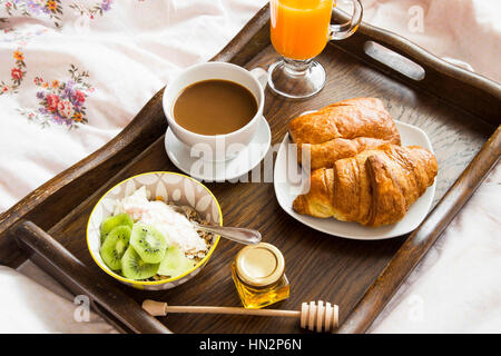 Breakfast in bed in wooden tray with coffee, juice, muesli with yogurt, croissants Stock Photo