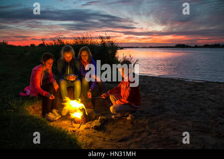 Mother and three children roasting marshmallows in campfire on riverbank at sunset Stock Photo