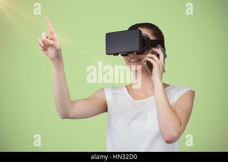 Beautiful woman gesturing while suing virtual reality headset and talking mobile phone against green background Stock Photo