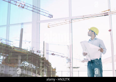 Crane and building construction site against architect in yellow hard hat with plans looking up at window Stock Photo