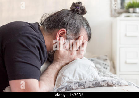 Man covering his face in grief, lying on a bed in bedroom Stock Photo