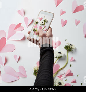 Female hand making photos of white background decoration in Valentine's Day. Mobile phone's camera making pictures of hearts. Valentine's Day concept. Stock Photo