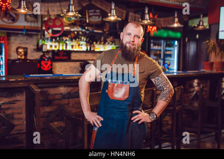 Bearded barman with tattoos and watches wearing an apron standing near the bar. Stock Photo