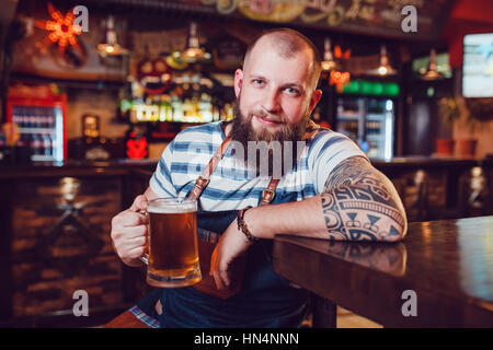 Bearded barman with tattoos wearing an apron sitting in bar and holding a glass of beer Stock Photo