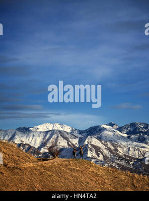 Two young girls stand on a hiking trail overlooking the Wasatch mountains of Salt Lake City, Utah. The mountain peaks are snow-covered and dramatic. Stock Photo