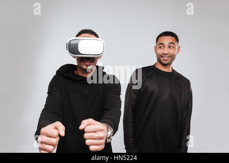 Image of two joyful african american young men standing and laughing while using 3d virtual reality device. Stock Photo