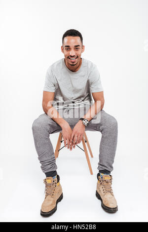 Picture of attractive young dark skinned man sitting over white background and looking at camera. Stock Photo