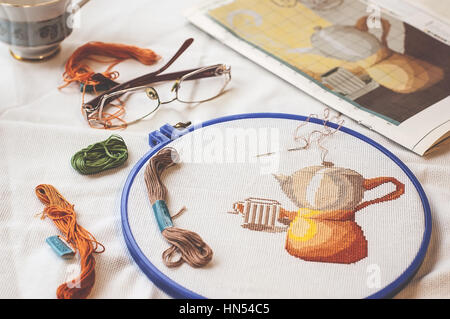 Cross-stitch set : hoop with embroidered teapot pattern, glasses, scheme, scissors, canvas and colorful yarn. Freelance, hobby, handmade concept Stock Photo