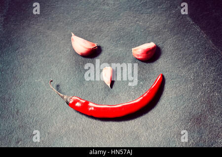 Smiling face made of red chili pepper and cloves of garlic on slate background, conceptual picture, selective focus. Stock Photo