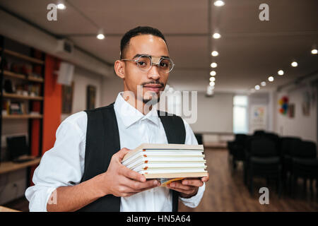 Image of young attractive african businessman wearing eyeglasses standing in library while holding notebooks. Look at camera. Stock Photo