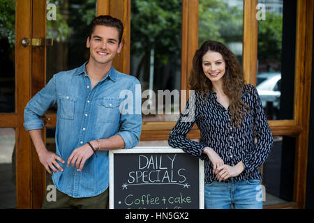 Portrait of smiling man and woman leaning on menu board outside the cafe Stock Photo
