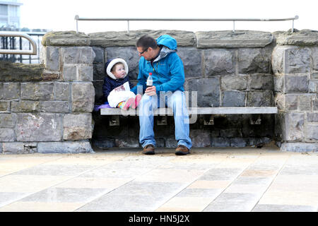 A grandfather sitting outdoors on a wooden bench sharing a bag of traditional fish and chips with his young granddaughter Stock Photo