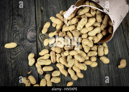 Spilling from white metal bucket of peanuts in the shell on a gray wooden surface, vintage toning Stock Photo