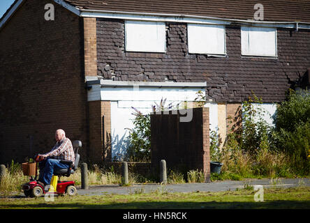 Old overweight fat man on mobility scooter passing old housing stock run down boarded up windows and empty in the East Manchester clearance of run dow Stock Photo