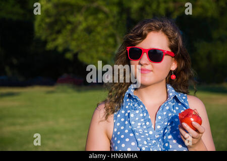 Young female with red sunglasses and blue polka dot holding a whole tomato in park Stock Photo