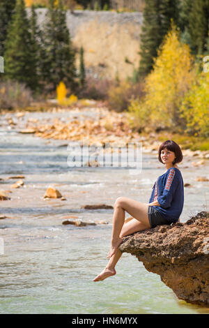 White water Mineral Creek stream in Colorado, USA during the fall with golden aspens and woman on edge dipping feet in water Stock Photo