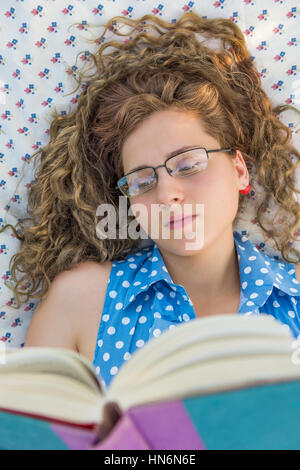 Young woman with glasses lying down reading book on blanket with curly hair from above