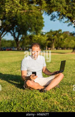 Young man looking at phone and smiling sitting outside in green grass park with laptop Stock Photo