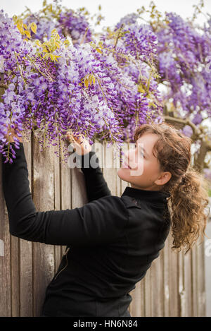 Young woman hanging onto wooden fence looking at smelling lilac purple wisteria flowers Stock Photo