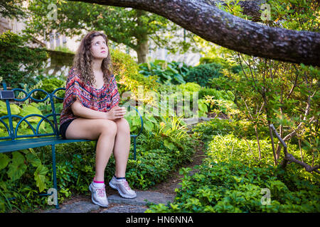 Young sad woman sitting on bench in garden looking up praying Stock Photo