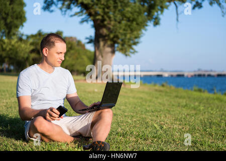 Young man looking at laptop and holding phone, sitting outside in green grass park Stock Photo
