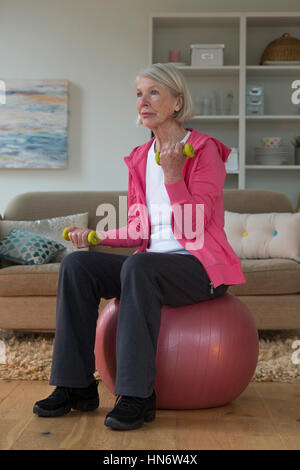 Senior lady sitting on a gym ball in her home whilst using dumbbells. Stock Photo