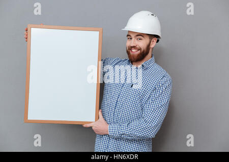 Image of happy builder holding copyspace board standing over grey background. Stock Photo