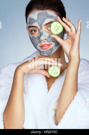 Beautiful girl in white bathrobe and with mask of clay. Photo of brunette woman holding cucumber slices near her face on blue background. Grooming him Stock Photo