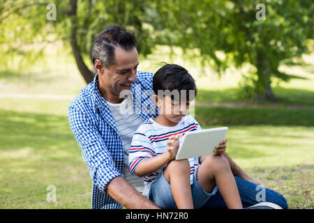 Boy sitting on his fathers lap and using digital tablet in park on a sunny day Stock Photo