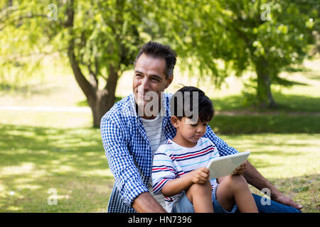 Boy sitting on his fathers lap and using digital tablet in park on a sunny day Stock Photo