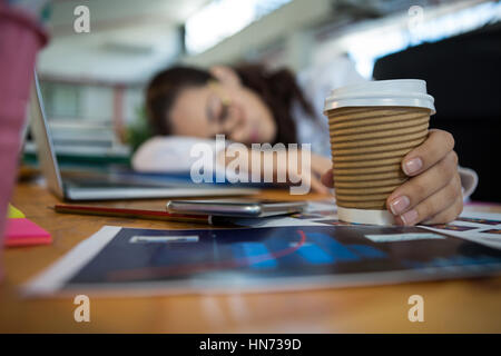 Tired female graphic designer holding disposable cup while sleeping on desk in office Stock Photo