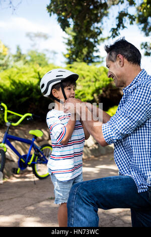 Father assisting son in wearing bicycle helmet in park on a sunny day Stock Photo