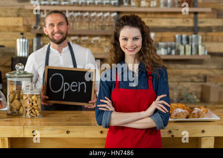 Portrait of waitress standing with arms crossed at counter in cafÃƒÂ© with waiter holding open sign board in background Stock Photo