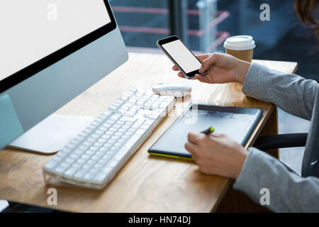Female graphic designer using graphics tablet and mobile phone at desk in office Stock Photo