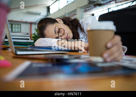 Tired female graphic designer holding disposable cup while sleeping on desk in office Stock Photo