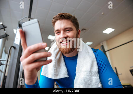 Photo of smiling young sportsman sitting in gym and looking at phone. Focus on man. Stock Photo