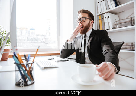 Image of handsome bearded businessman sitting in office while holding coffee and yawning Stock Photo