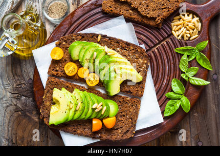 sandwich with rye bread on a board for cutting: avocado, yellow tomatoes, pine nuts and basil leaves. grated black pepper and olive oil. on the old wo Stock Photo