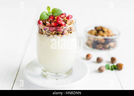 Healthy breakfast concept. Vanilla cream with fresh granola in a glass topped with pomegranate and mint arranged on white wooden table. Stock Photo