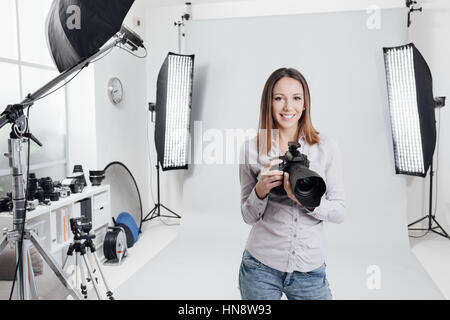 Young female photographer posing in the photo studio, she is smiling and holding a professional digital camera Stock Photo