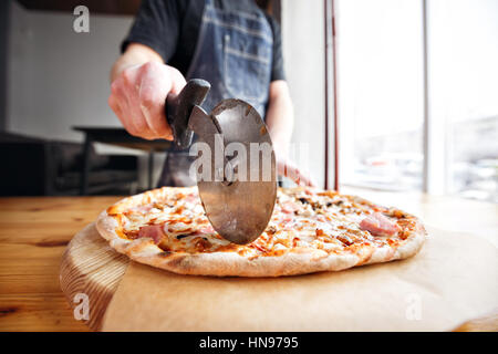 Closeup hand of chef baker in uniform blue apron cutting pizza at kitchen Stock Photo