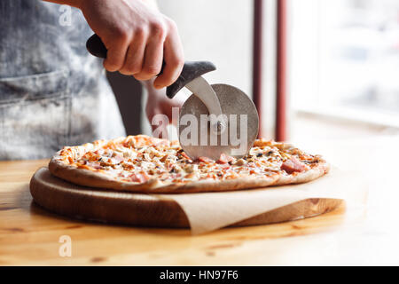 Closeup hand of chef baker in uniform blue apron cutting pizza at kitchen Stock Photo