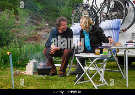 couple sitting in camping chairs in front of their camper and they are cooking on the barbecue Stock Photo